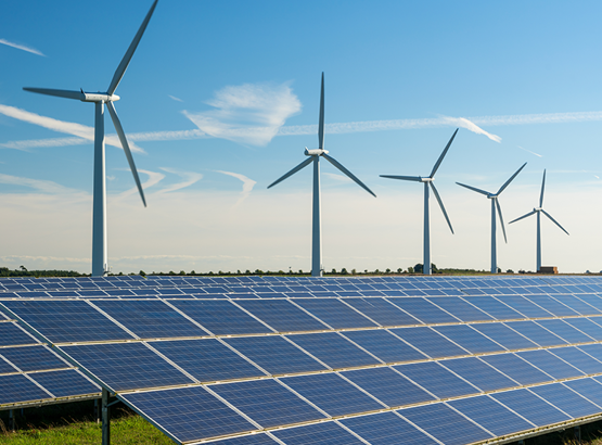 Rows of blue solar panels in the foreground with several tall wind turbines turning under a clear blue sky