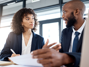 Two employees seated at a desk discussing documents in an office setting.