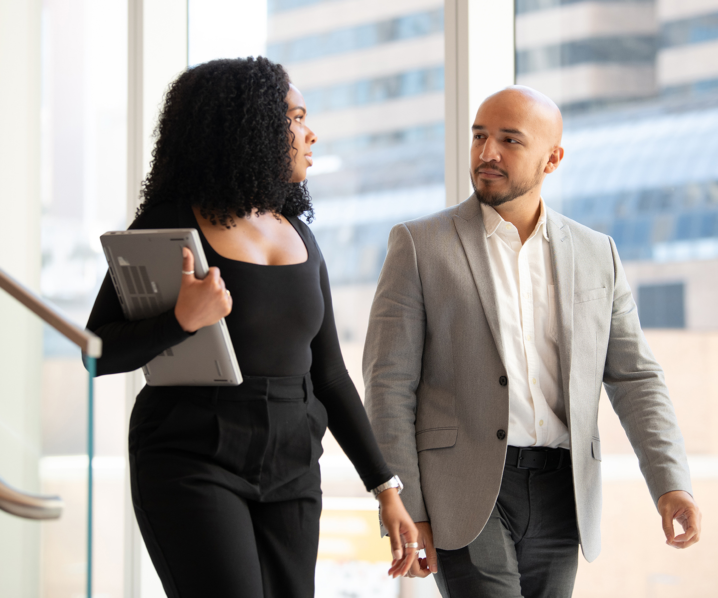 Two professionals walk side by side through a bright office hallway, one carrying a tablet, with city buildings visible through large windows